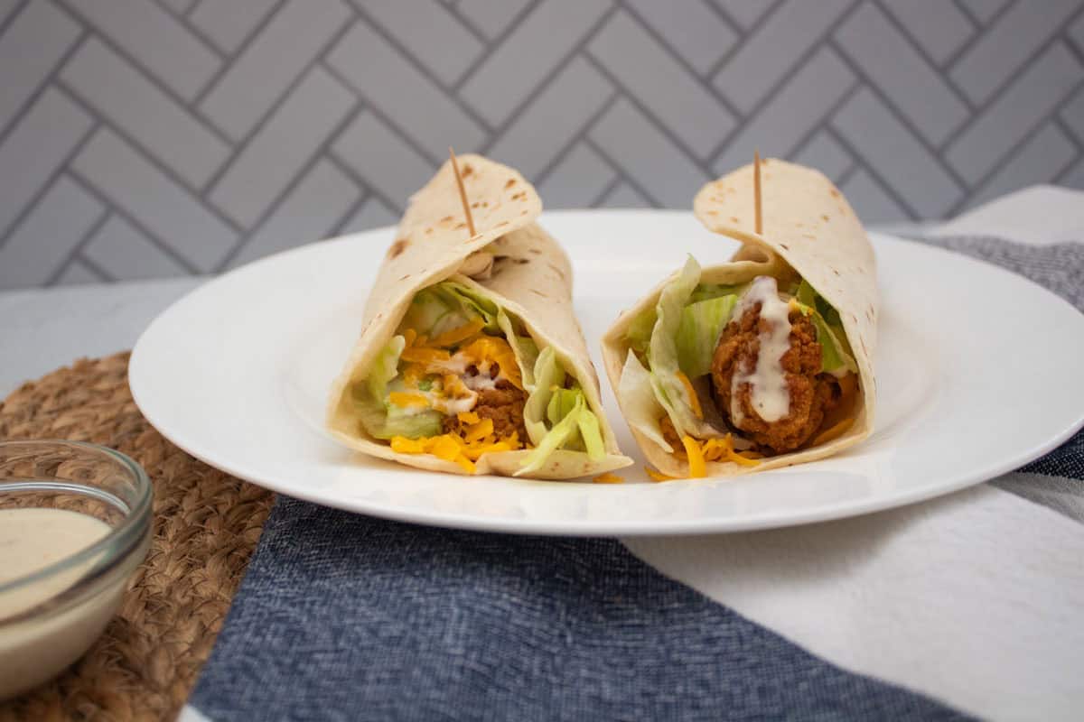 Two chicken wraps filled with lettuce, shredded cheese, and drizzled with creamy sauce, served on a white plate with toothpicks, placed on a blue and white cloth. A bowl of sauce is visible on the side.