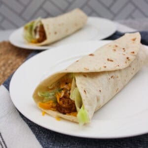 A close-up of a flour tortilla wrap filled with shredded cheese, lettuce, and chicken on a white plate, with a second wrap and plate blurred in the background.