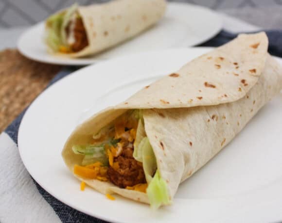 A close-up of a flour tortilla wrap filled with shredded cheese, lettuce, and chicken on a white plate, with a second wrap and plate blurred in the background.