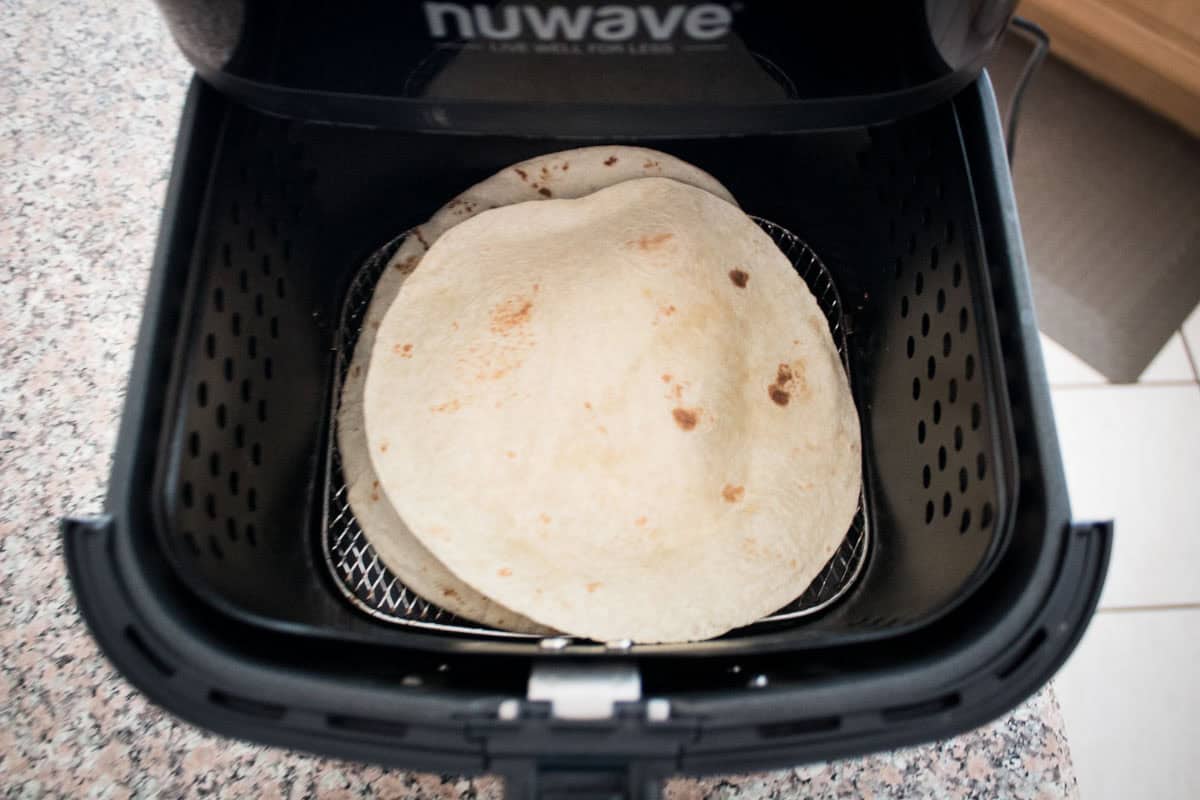Two flour tortillas are placed inside the basket of a black air fryer on a speckled countertop, ready to be warmed.