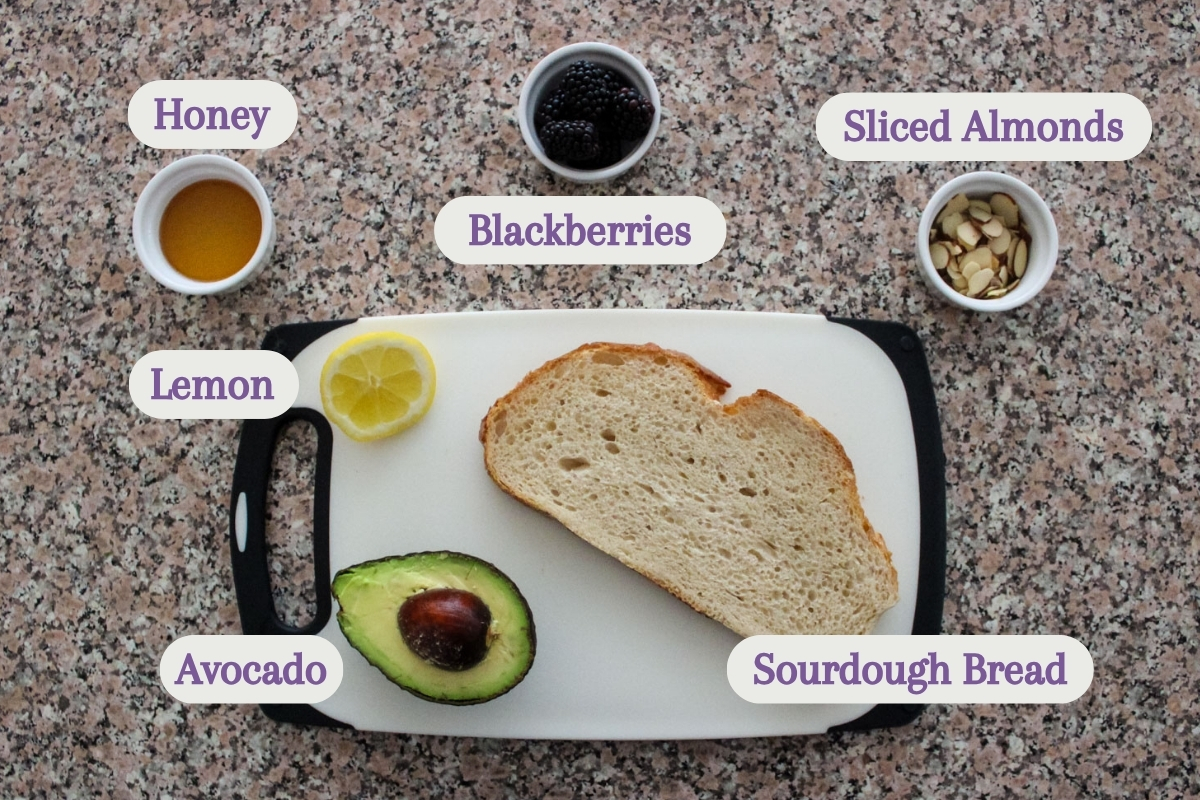 A cutting board with half an avocado, a lemon wedge, a sourdough bread slice, and small bowls of honey, blackberries, and sliced almonds labeled on a speckled countertop.