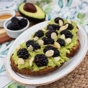 A slice of toast topped with mashed avocado, blackberries, and almond slices on a white plate. In the background, there is half an avocado, a small bowl of blackberries, and a small dish of honey.