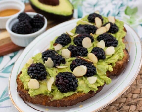 A slice of toast topped with mashed avocado, blackberries, and almond slices on a white plate. In the background, there is half an avocado, a small bowl of blackberries, and a small dish of honey.