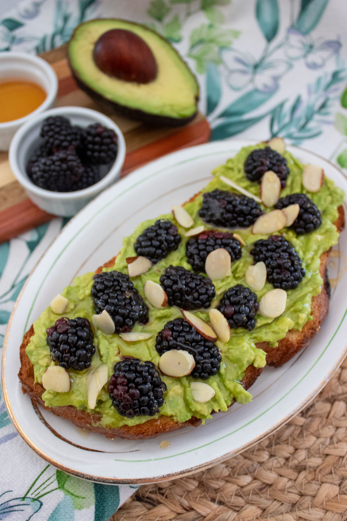A slice of toast topped with mashed avocado, blackberries, and almond slices on a white plate. In the background are a halved avocado, honey, and a small bowl of blackberries.