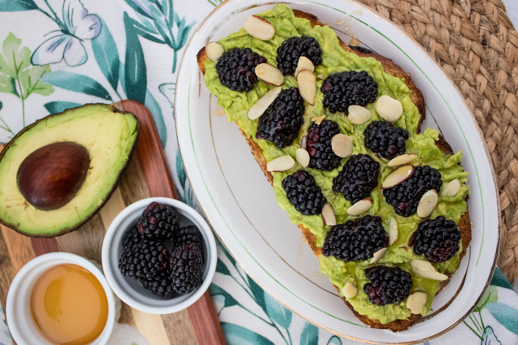 A plate with avocado toast topped with blackberries and sliced almonds. Next to it are a halved avocado, a bowl of blackberries, and a small dish of honey on a patterned tablecloth.