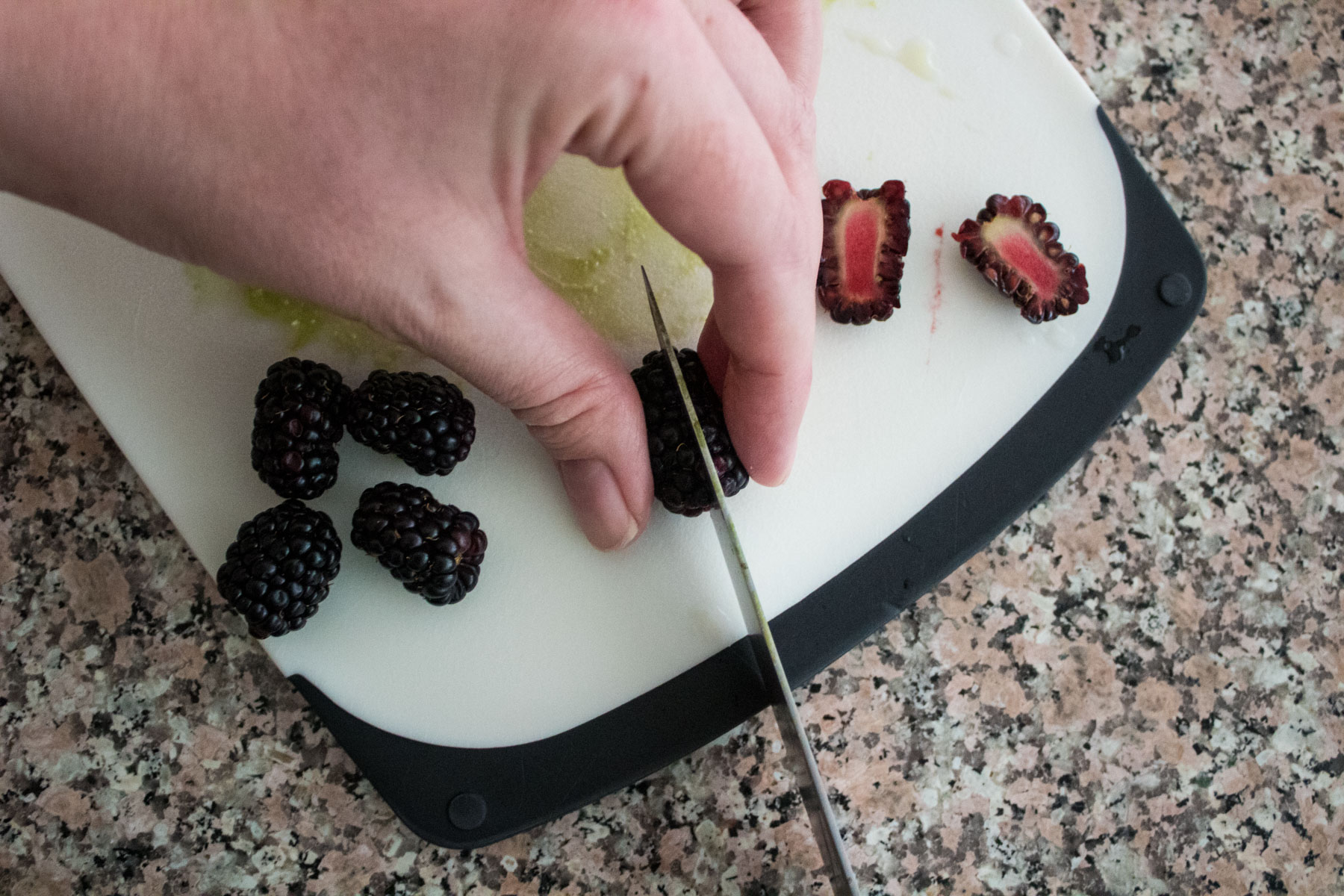 A hand slices blackberries on a white cutting board with a knife. Several whole and halved blackberries are visible on a granite countertop.