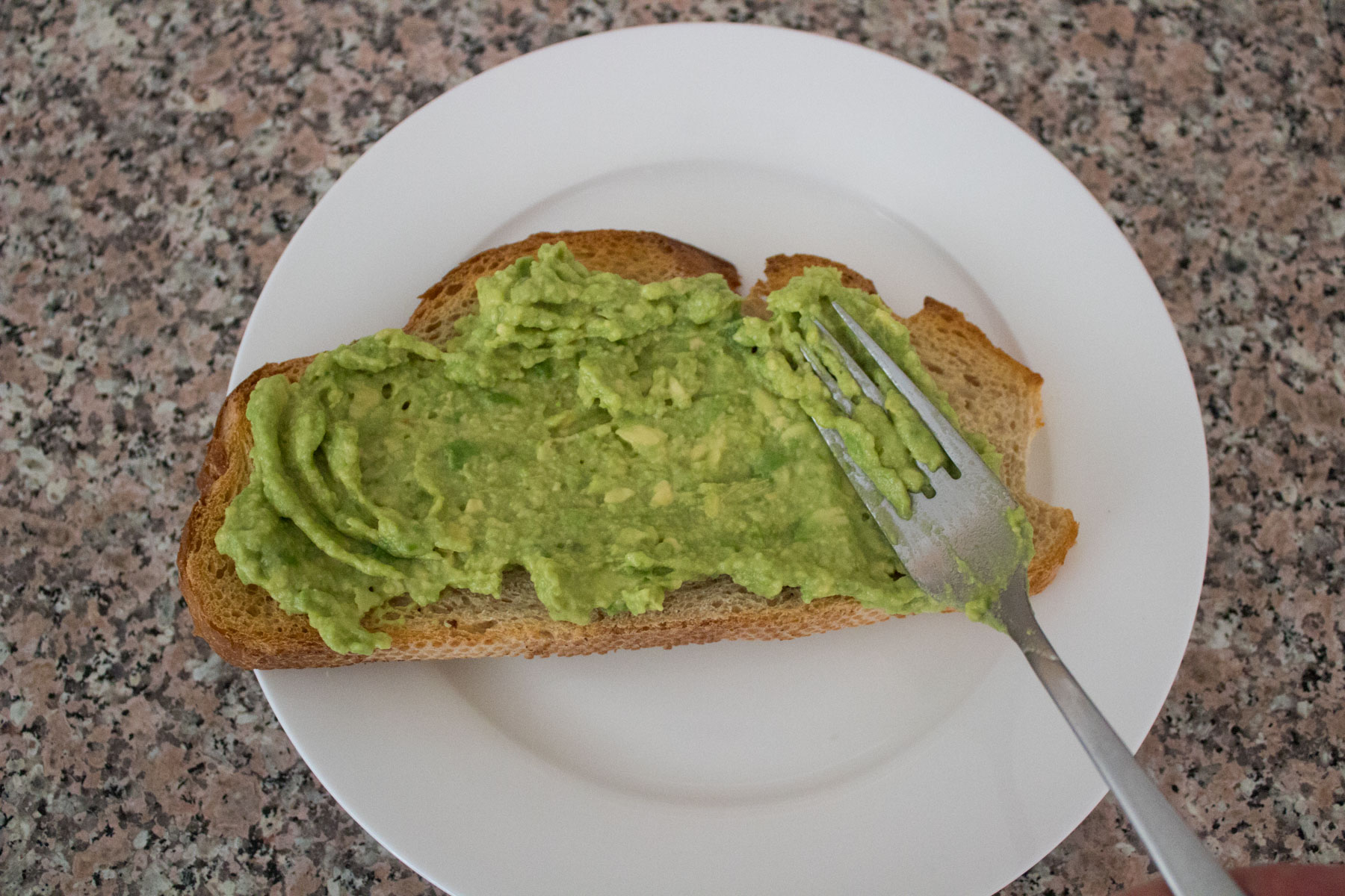 A slice of toast on a white plate is being spread with mashed avocado using a fork, set on a speckled countertop.