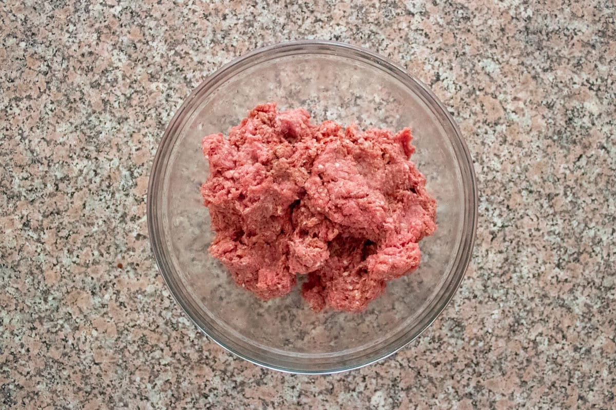 A clear glass bowl filled with raw ground beef is placed on a speckled kitchen countertop, viewed from above.