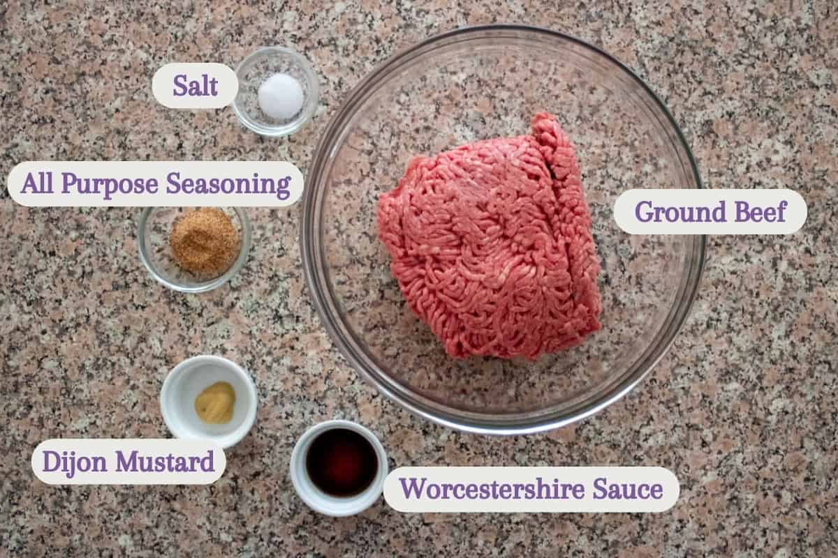 A glass bowl of ground beef sits on a counter, surrounded by small bowls containing salt, all purpose seasoning, Dijon mustard, and Worcestershire sauce, each labeled with text.