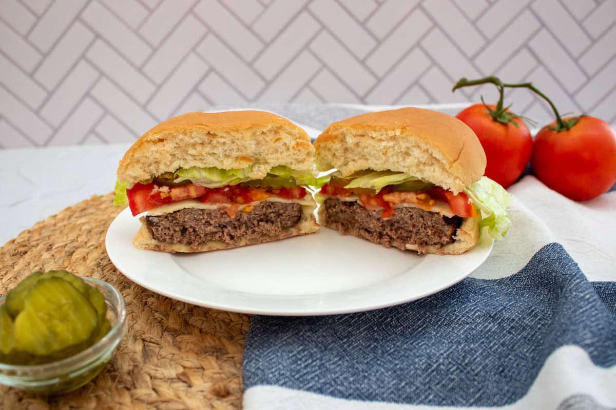A hamburger cut in half sits on a white plate, showing lettuce, tomato, pickles, and beef patty. Tomatoes and a small bowl of pickles are nearby on a woven mat with a blue and white cloth.