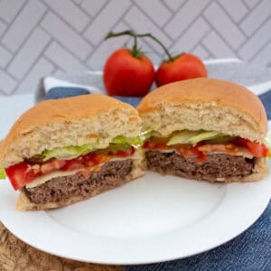 A hamburger cut in half on a white plate, showing layers of lettuce, tomato, cheese, and beef patty. Two whole tomatoes and a blue napkin are in the background.