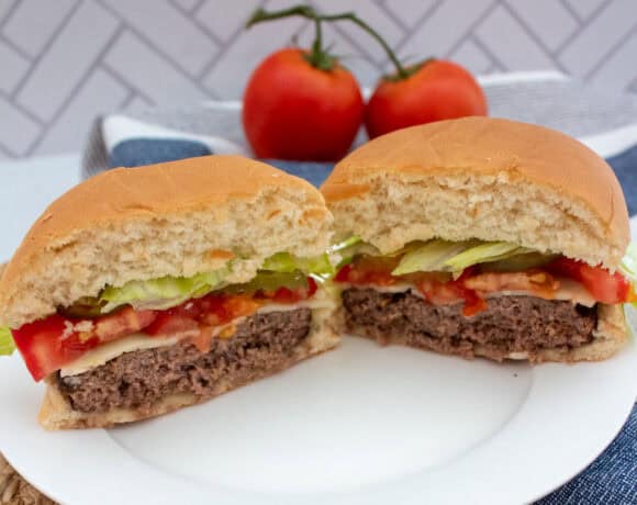 A hamburger cut in half on a white plate, showing layers of lettuce, tomato, cheese, and beef patty. Two whole tomatoes and a blue napkin are in the background.