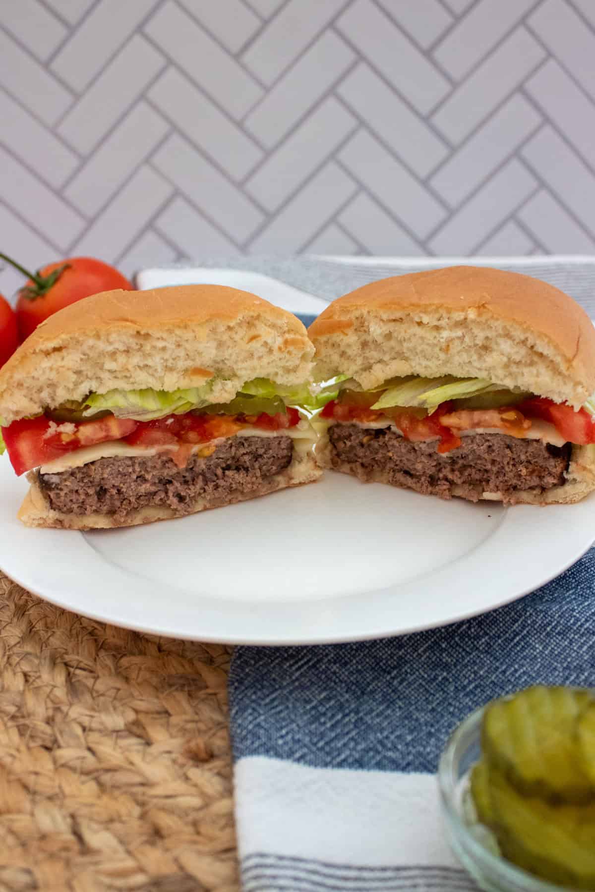 A hamburger cut in half on a white plate, showing layers of beef patty, lettuce, tomato, pickles, and sauce in a soft bun. Tomatoes and a bowl of pickles are in the background.