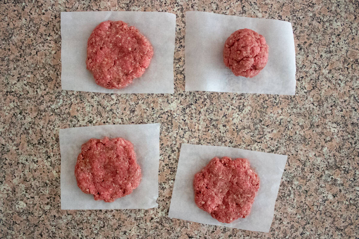 Four portions of raw ground beef, three flattened and one in a ball, are placed on separate pieces of parchment paper on a speckled countertop.