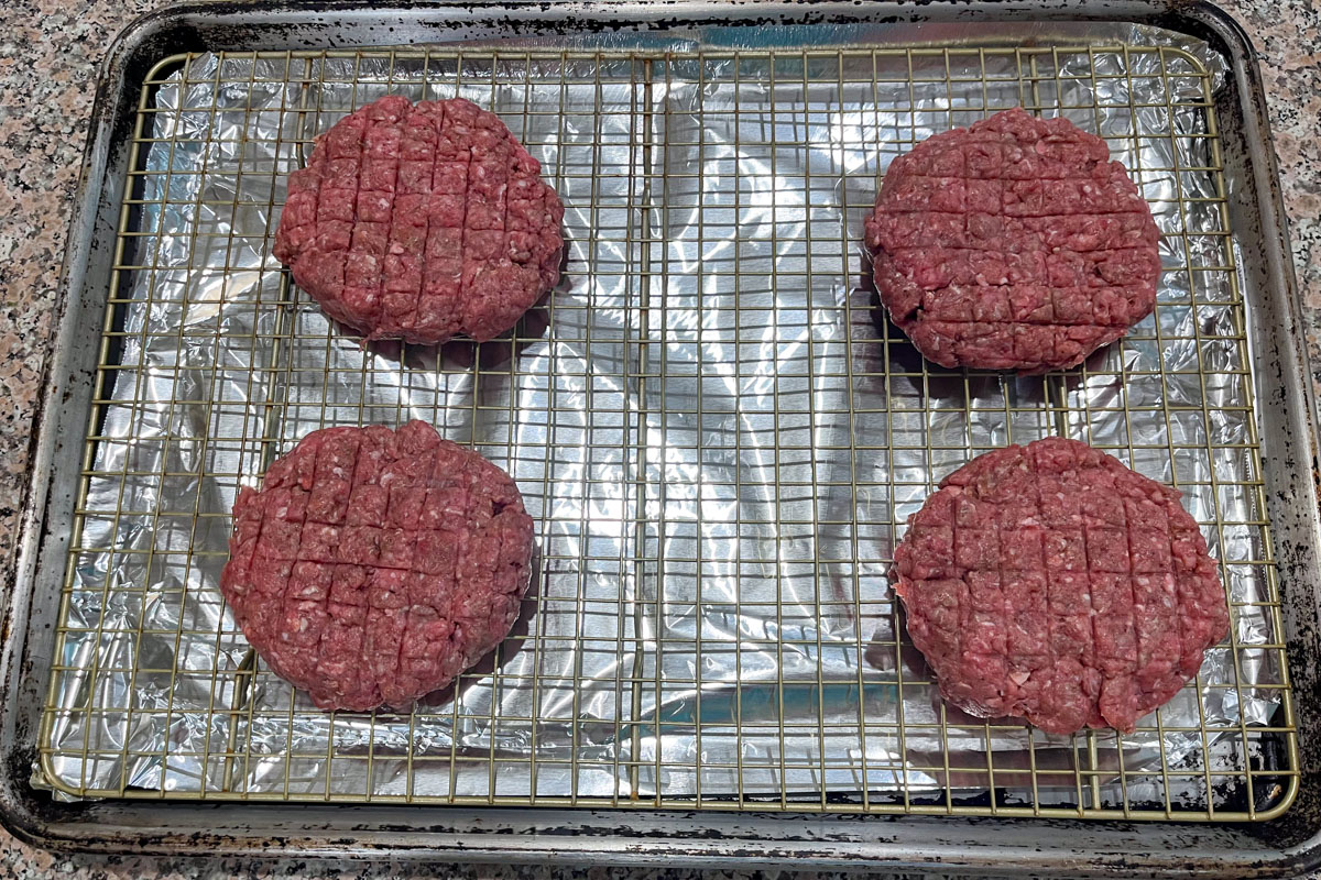 Four raw beef burger patties with a crisscross pattern are placed on a wire rack over a foil-lined baking sheet, ready for cooking on a kitchen countertop.