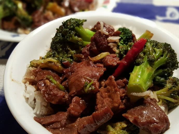 Beef and broccoli stir-fry served over white rice in a white bowl, with a dried red chili pepper visible on top.