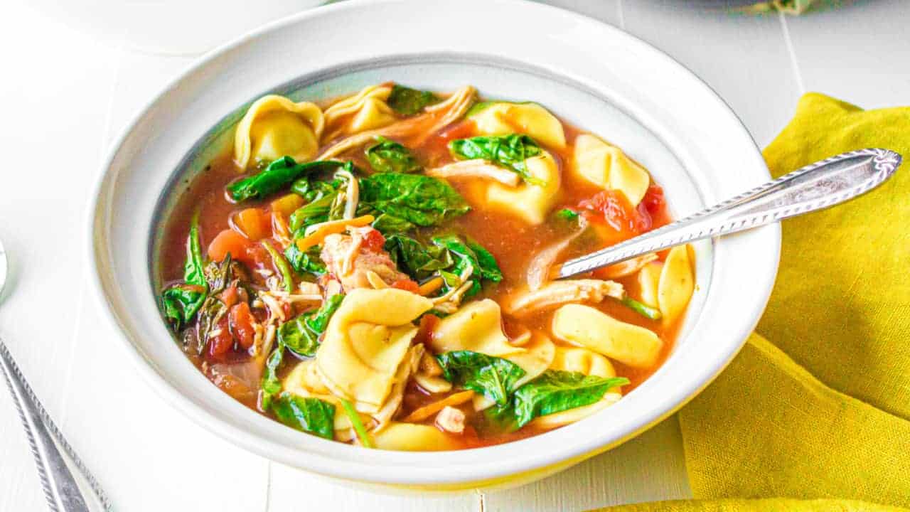A bowl of tortellini soup with spinach, shredded chicken, tomatoes, and broth. A silver spoon rests in the bowl, and a yellow napkin is nearby on a white table.