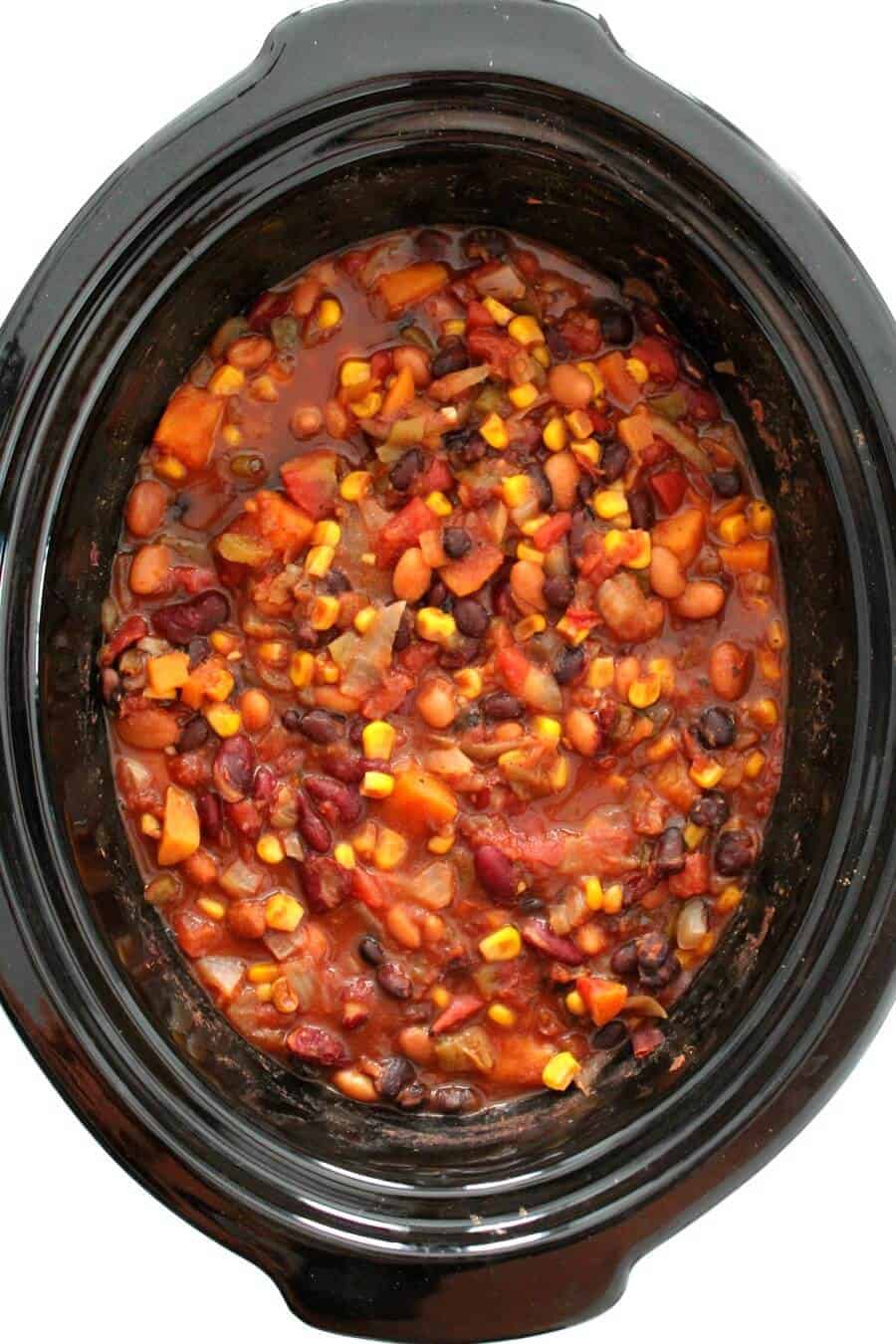 A slow cooker filled with vegetarian chili, containing beans, corn, diced tomatoes, and chunks of vegetables in a rich, red sauce.