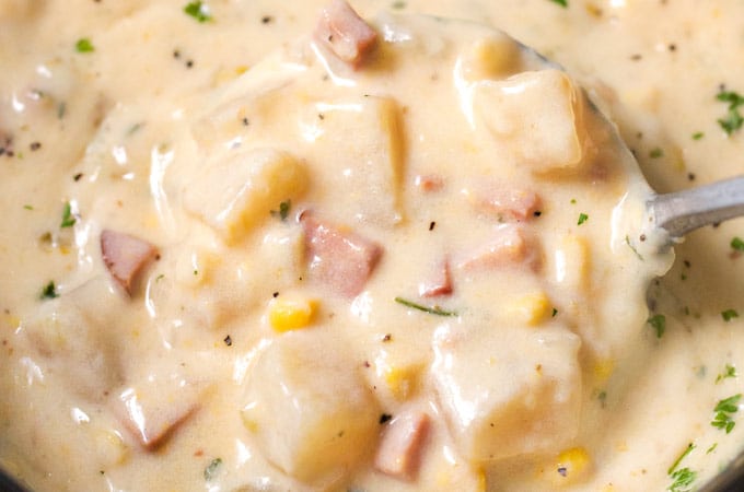 A close-up of creamy potato soup with diced ham, corn, herbs, and black pepper, being ladled from a pot.