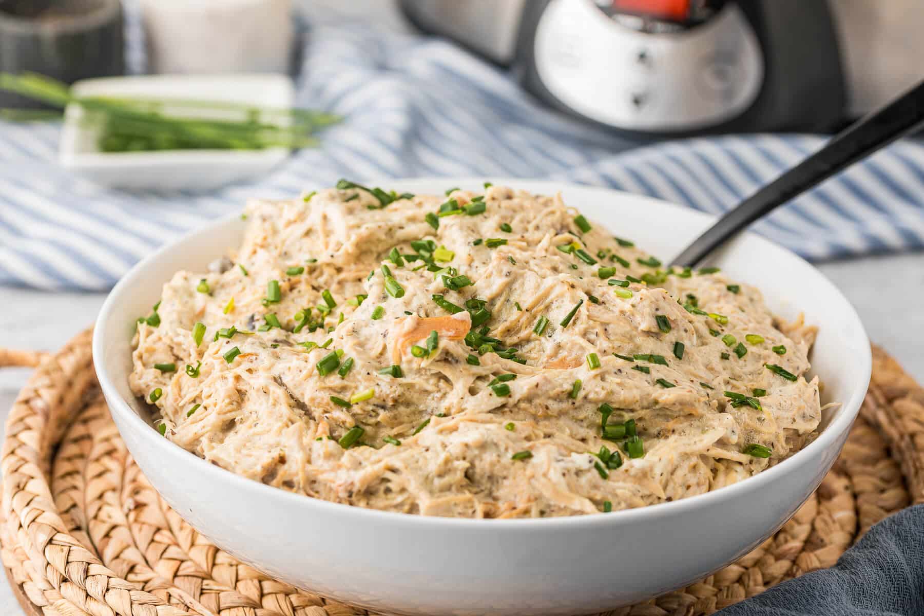 A white bowl filled with creamy shredded chicken mixed with herbs and chopped chives, placed on a woven placemat with a spoon inside. A striped cloth and kitchen items are blurred in the background.