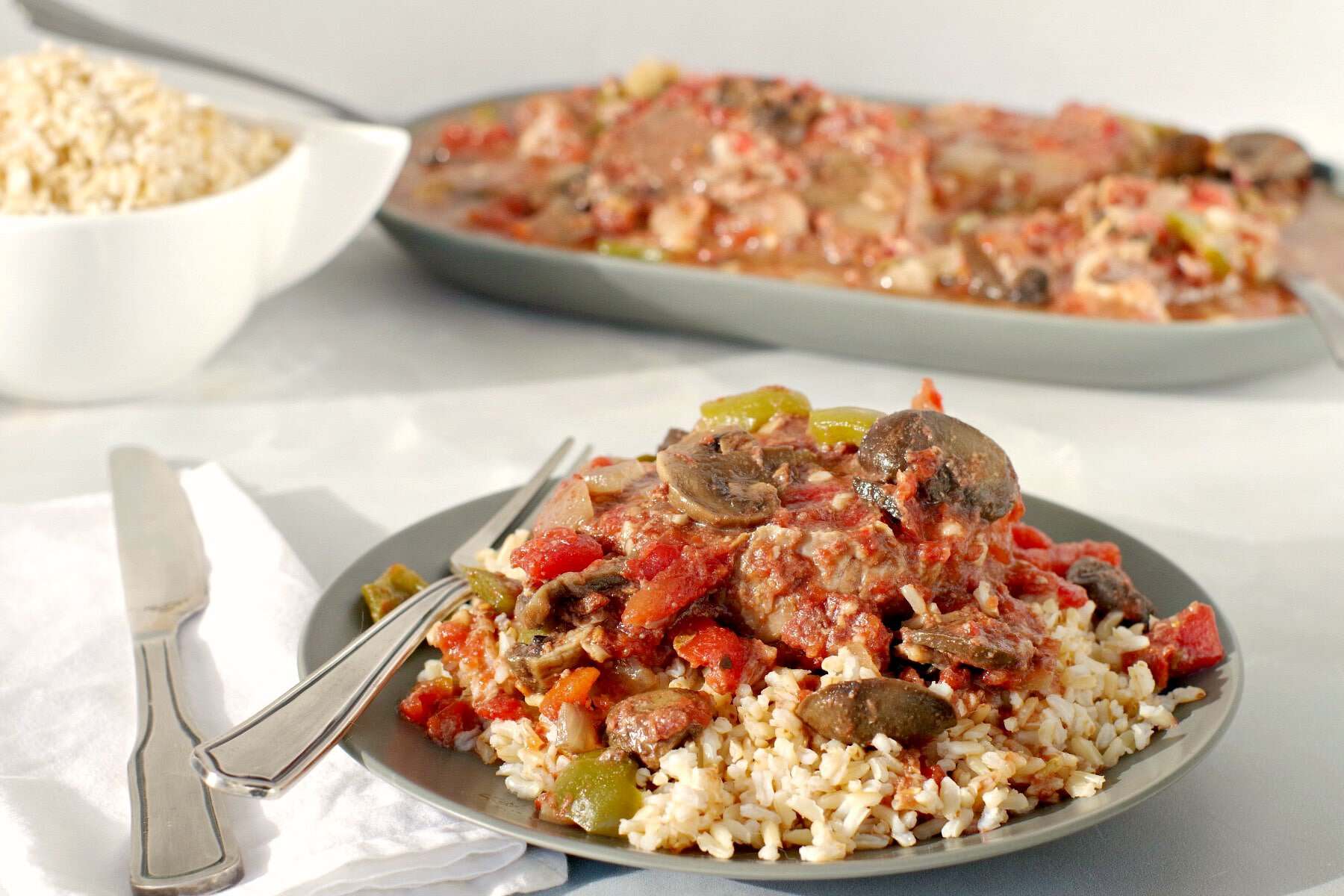 A plate of swiss steak with mushrooms, tomatoes, and peppers served over brown rice, with a fork and knife on the side.