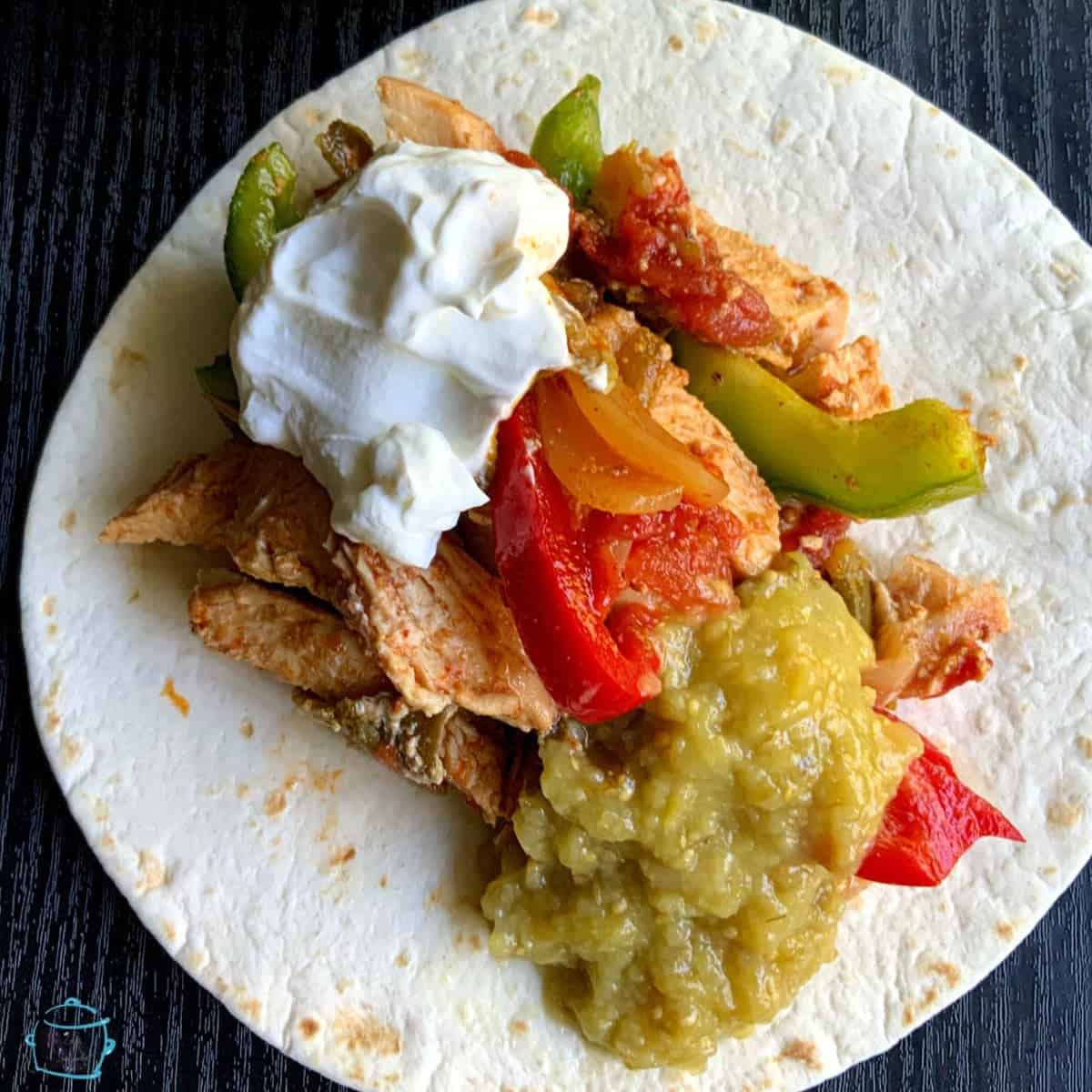 A flour tortilla topped with cooked chicken, sautéed bell peppers, onions, chunky green salsa, and a dollop of sour cream, all on a dark surface.