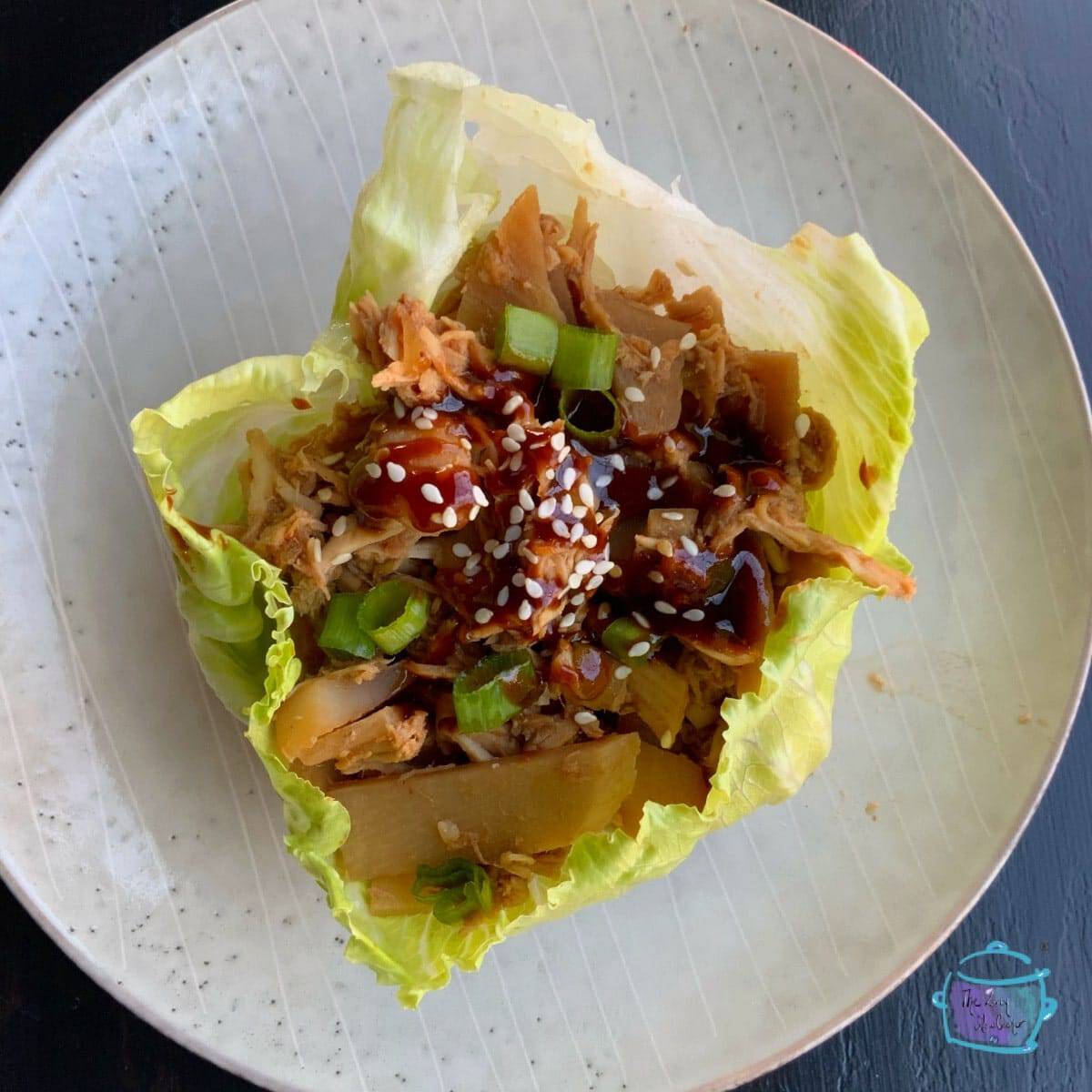 A single lettuce wrap filled with shredded meat, diced vegetables, green onions, sesame seeds, and a drizzle of dark sauce, served on a white plate with subtle stripes.