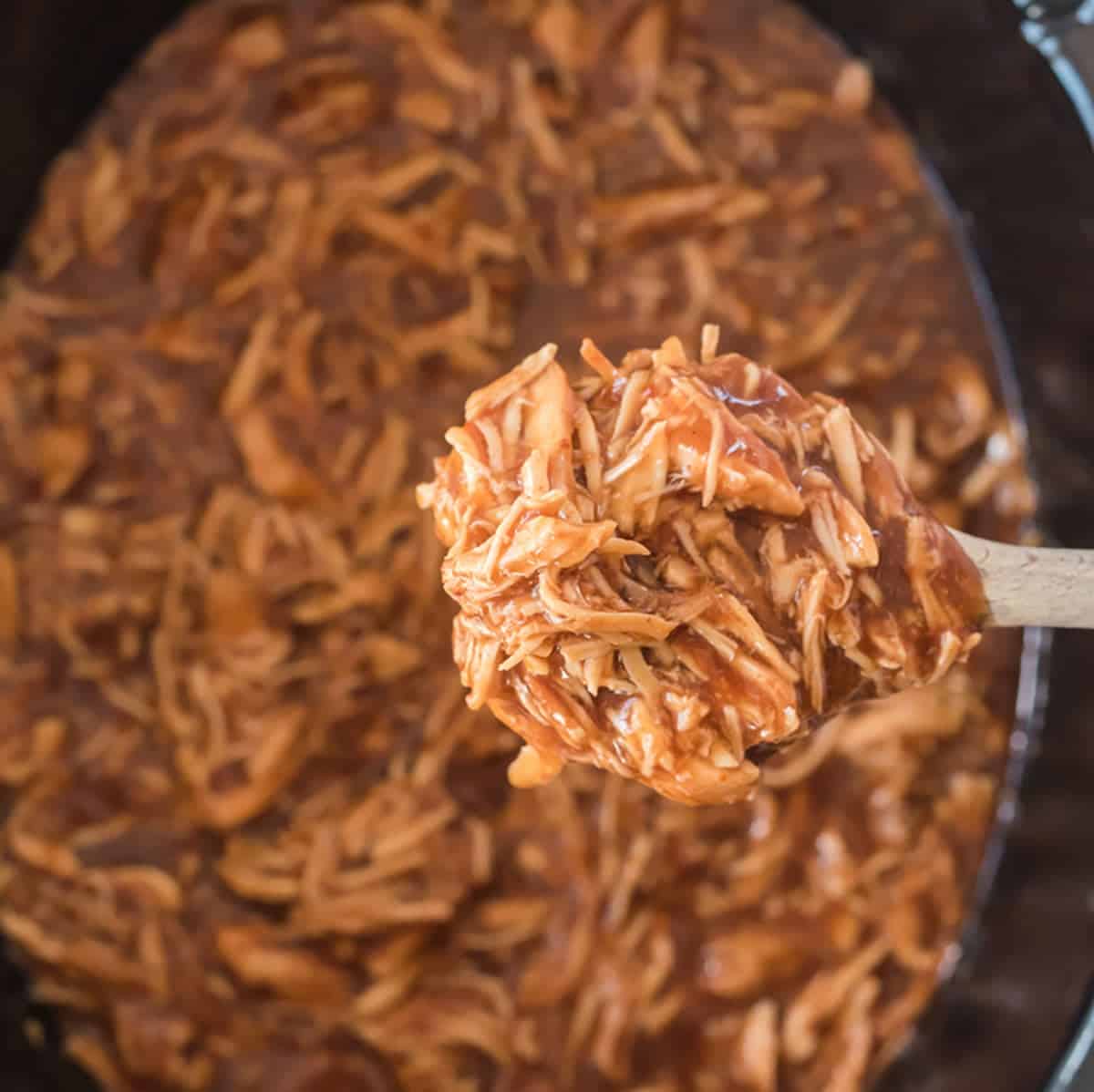 A wooden spoon holds up shredded barbecue chicken over a pot filled with more saucy shredded chicken.