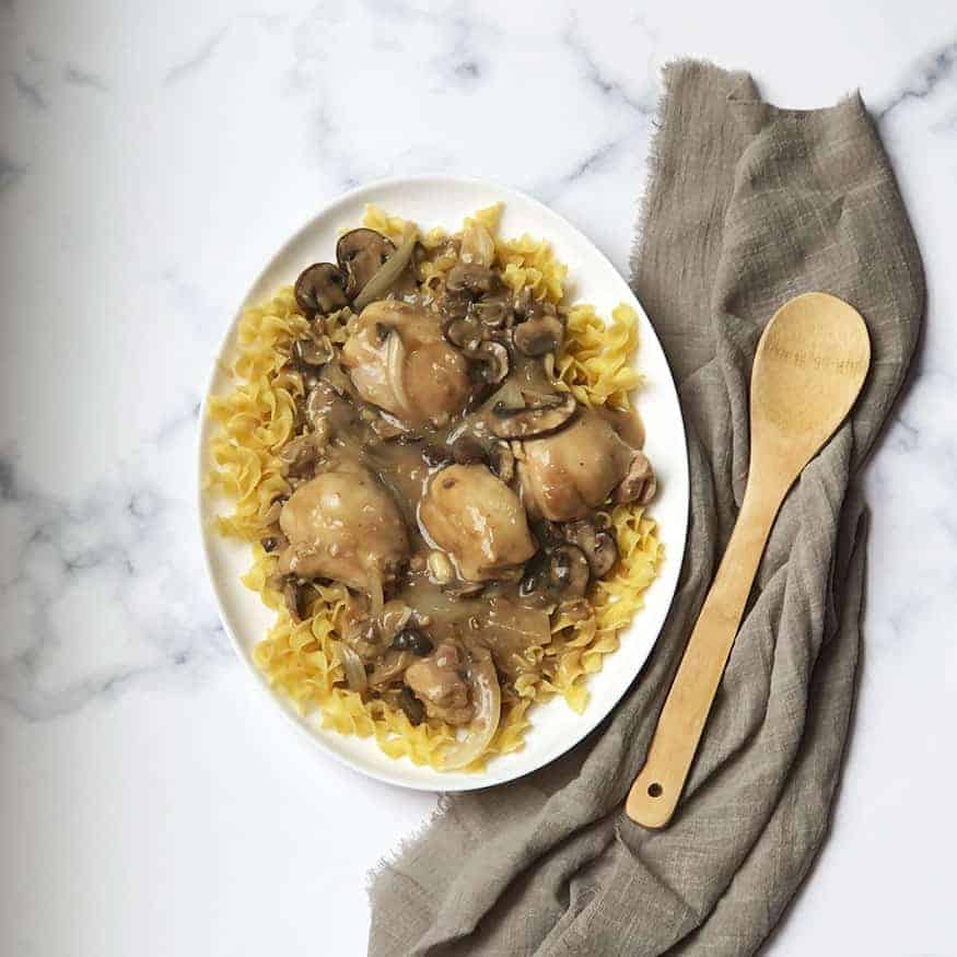 A white oval plate with egg noodles topped with chicken, mushrooms, and gravy. A wooden spoon and gray cloth napkin are placed beside the plate on a white marble surface.
