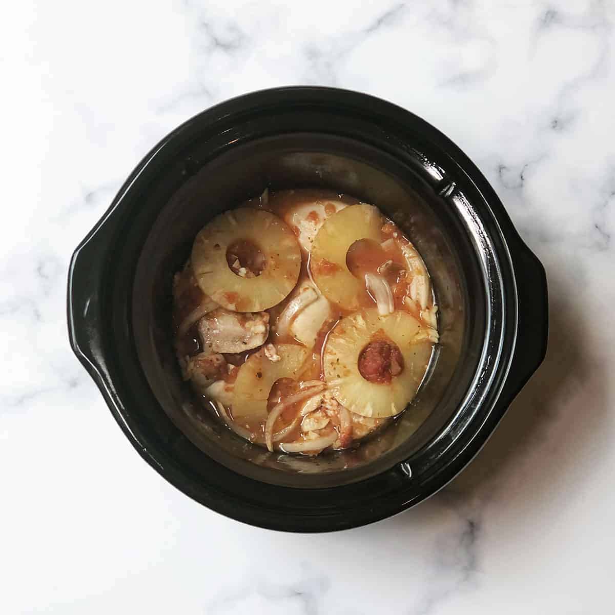 A black slow cooker filled with pieces of chicken, pineapple rings, and onions in sauce, sitting on a white marble surface.