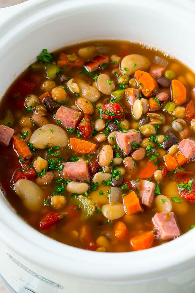A slow cooker filled with colorful bean and vegetable soup, featuring chunks of ham, carrots, celery, tomatoes, and beans, garnished with chopped parsley.
