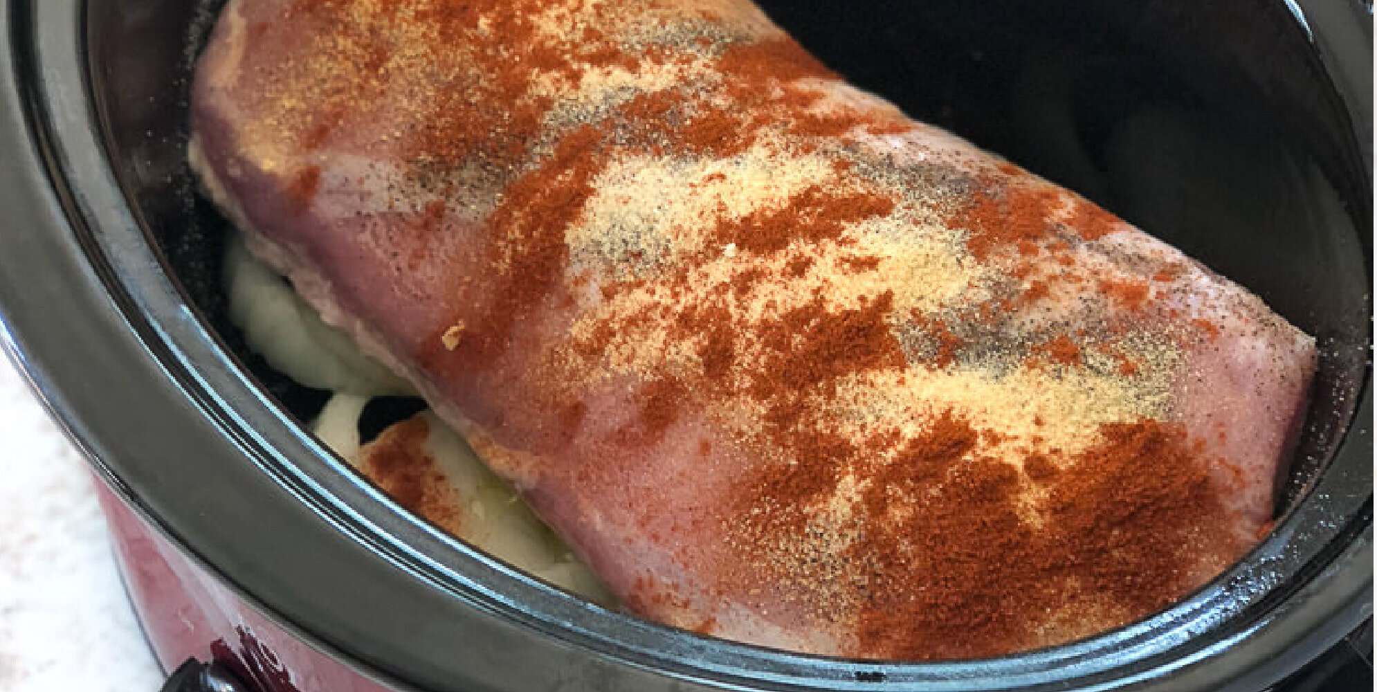 A raw pork roast seasoned with paprika and pepper in a slow cooker over sliced onions.