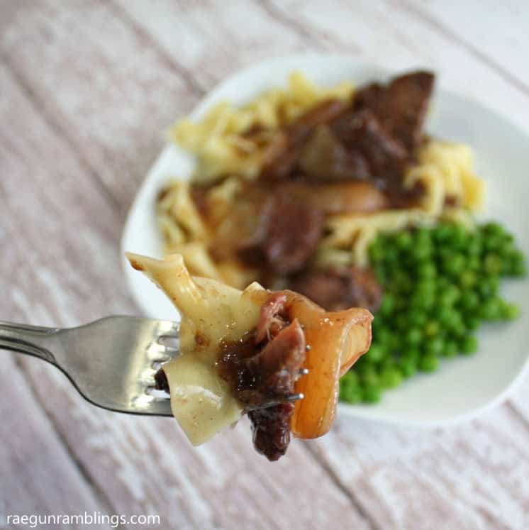 A fork holds a bite of beef, onions, and egg noodles above a plate with more beef, noodles, and a side of green peas.