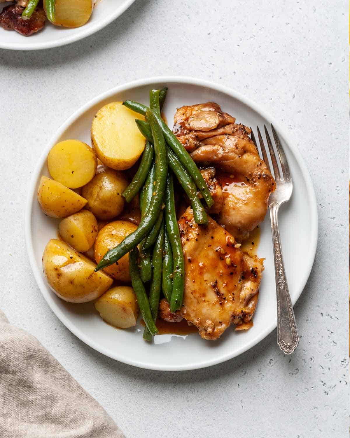 A white plate with glazed chicken thighs, roasted baby potatoes, and green beans. A fork rests on the side of the plate on a light surface.