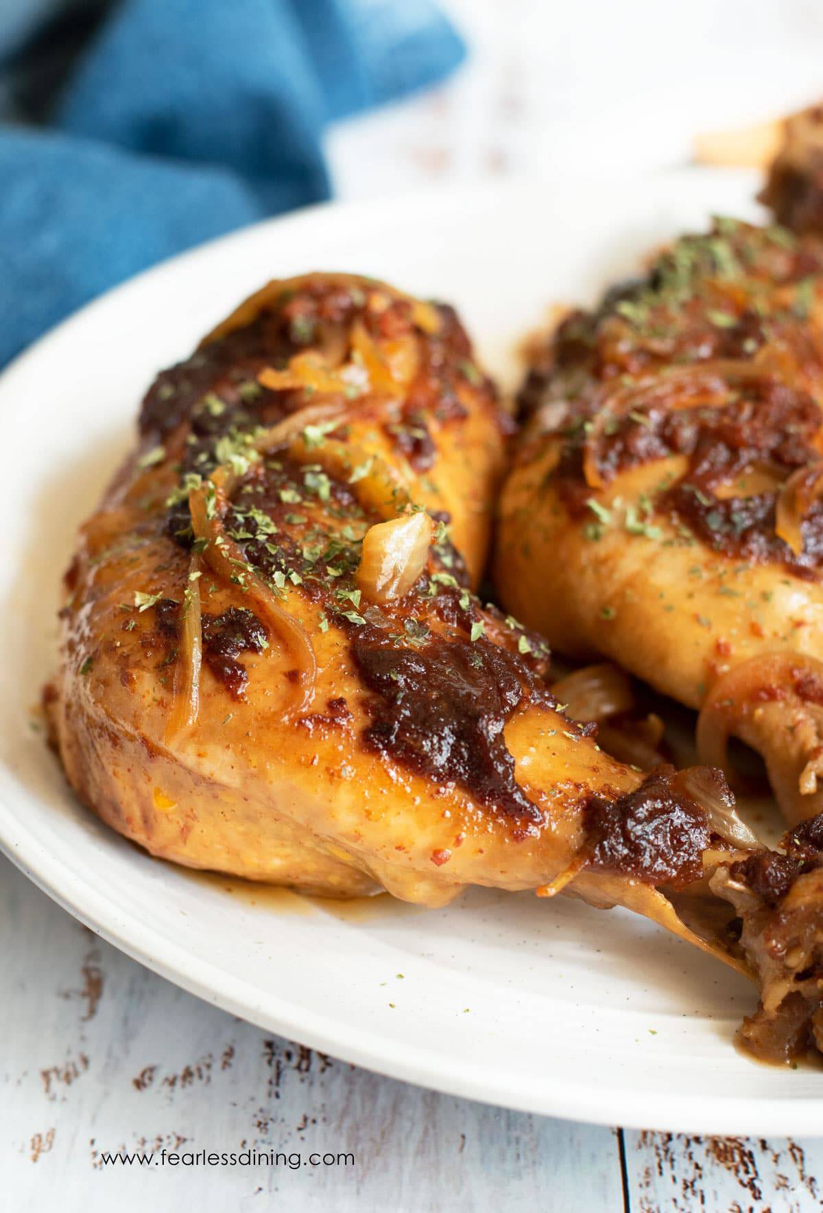 Two baked chicken leg quarters glazed with sauce and garnished with herbs are served on a white plate. The chicken appears juicy and flavorful with visible onion slices and a browned exterior.