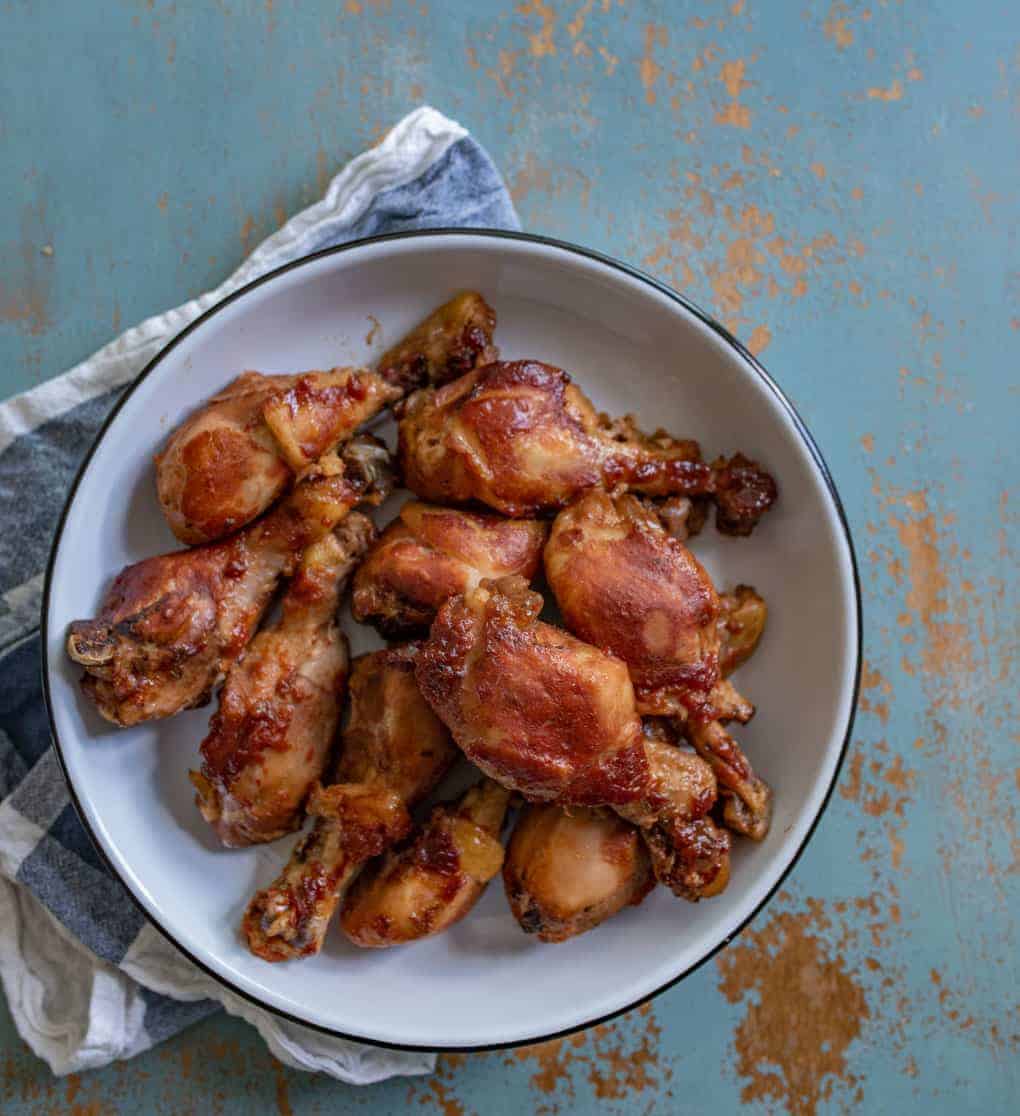 A white bowl filled with roasted chicken drumsticks sits on a blue and rust-colored surface, with a striped cloth partially visible underneath.