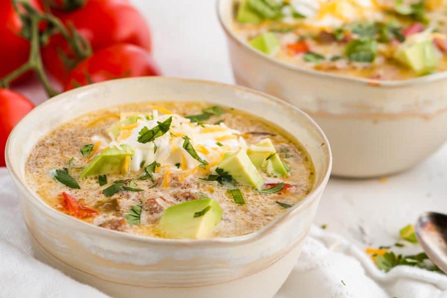A bowl of creamy soup topped with avocado, shredded cheese, sour cream, and cilantro, with tomatoes in the background.