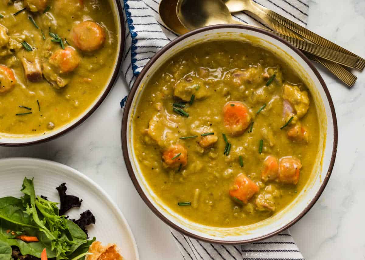 A bowl of hearty split pea soup with chunks of carrots and ham, garnished with herbs. A salad and toasted bread are partially visible on the side.