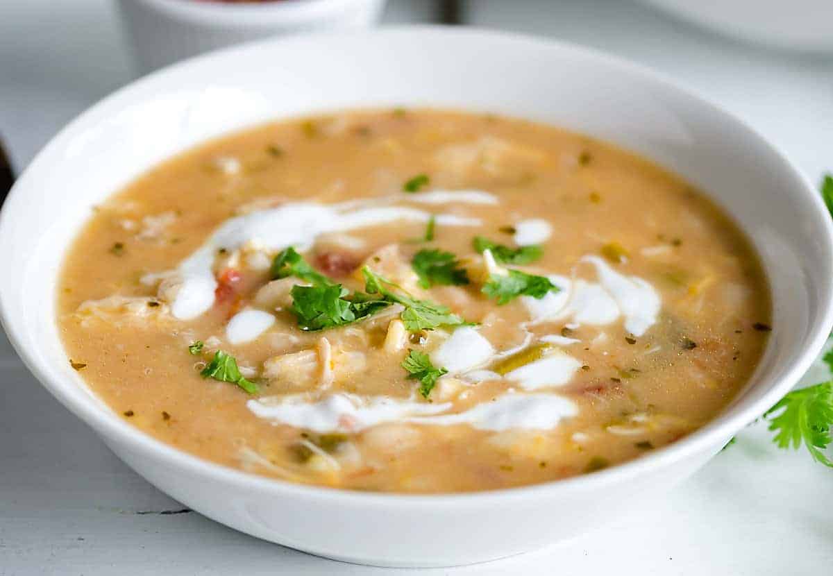 A bowl of creamy soup garnished with fresh cilantro and drizzled with sour cream, served in a white bowl on a white surface.